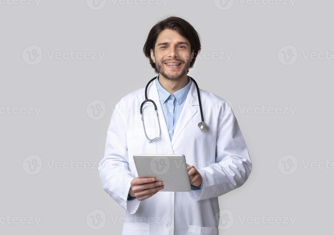 Medicine and technologies. Cheerful doctor using digital tablet in hands, grey studio background photo