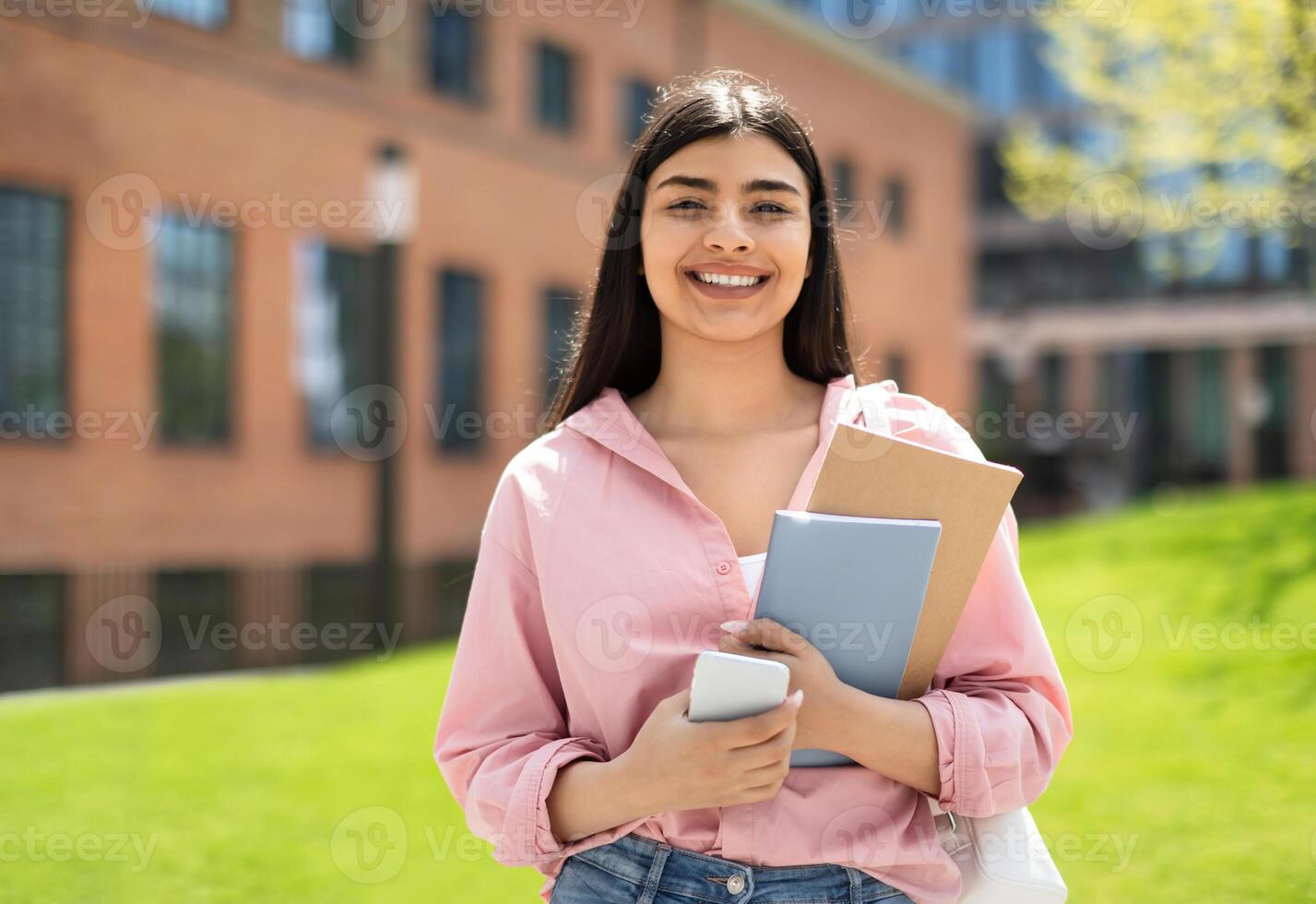 Portrait of happy student girl with cellphone and workbooks walking in college campus and smiling at camera, having break after classes outdoors, copy space photo