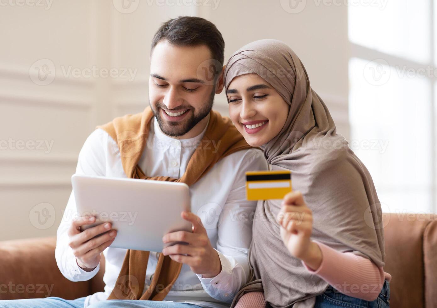 Online Shopping. Happy Muslim Couple Using Digital Tablet And Credit Card Buying And Purchasing Things Sitting On Sofa At Home. Joyful Customers, Modern Shopping And Sales Concept photo
