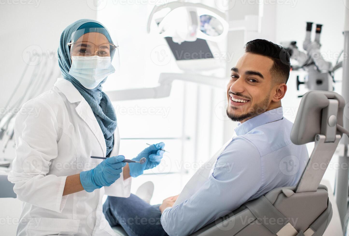 Portrait Of Happy Islamic Stomatologist Lady Having Check Up With Male Patient In Modern Dental Clinic, Muslim Dentist Woman In Hijab And Young Man Looking And Smiling At Camera, Closeup photo