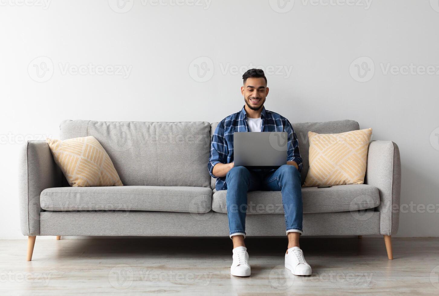 Remote Work. Handsome Middle Eastern Freelancer Guy Working With Laptop At Home, Arab Millennial Man Sitting On Couch In Living Room And Using Computer, Enjoying Distance Job Opportunities, Copy Space photo