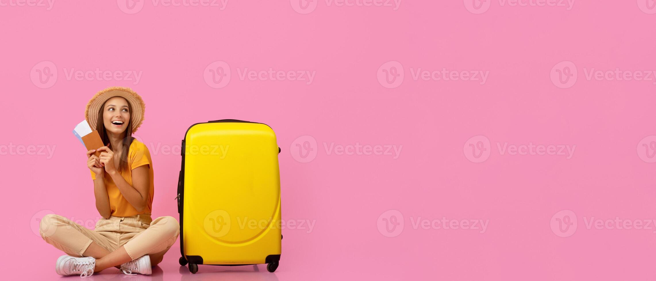 Surprised young woman traveler holding passport and flight tickets, sitting on floor next to suitcase and looking at copy space, pink background. Travelling deals and discounts, tourism concept photo