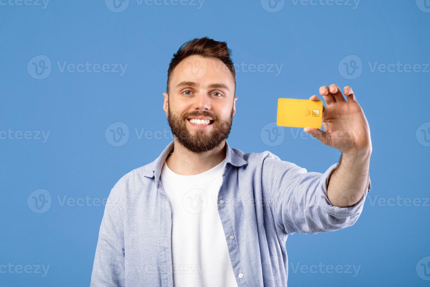 Positive millennial guy showing credit card and smiling on blue studio background. Young bearded man promoting contactless payment, using electronic money during coronavirus outbreak photo
