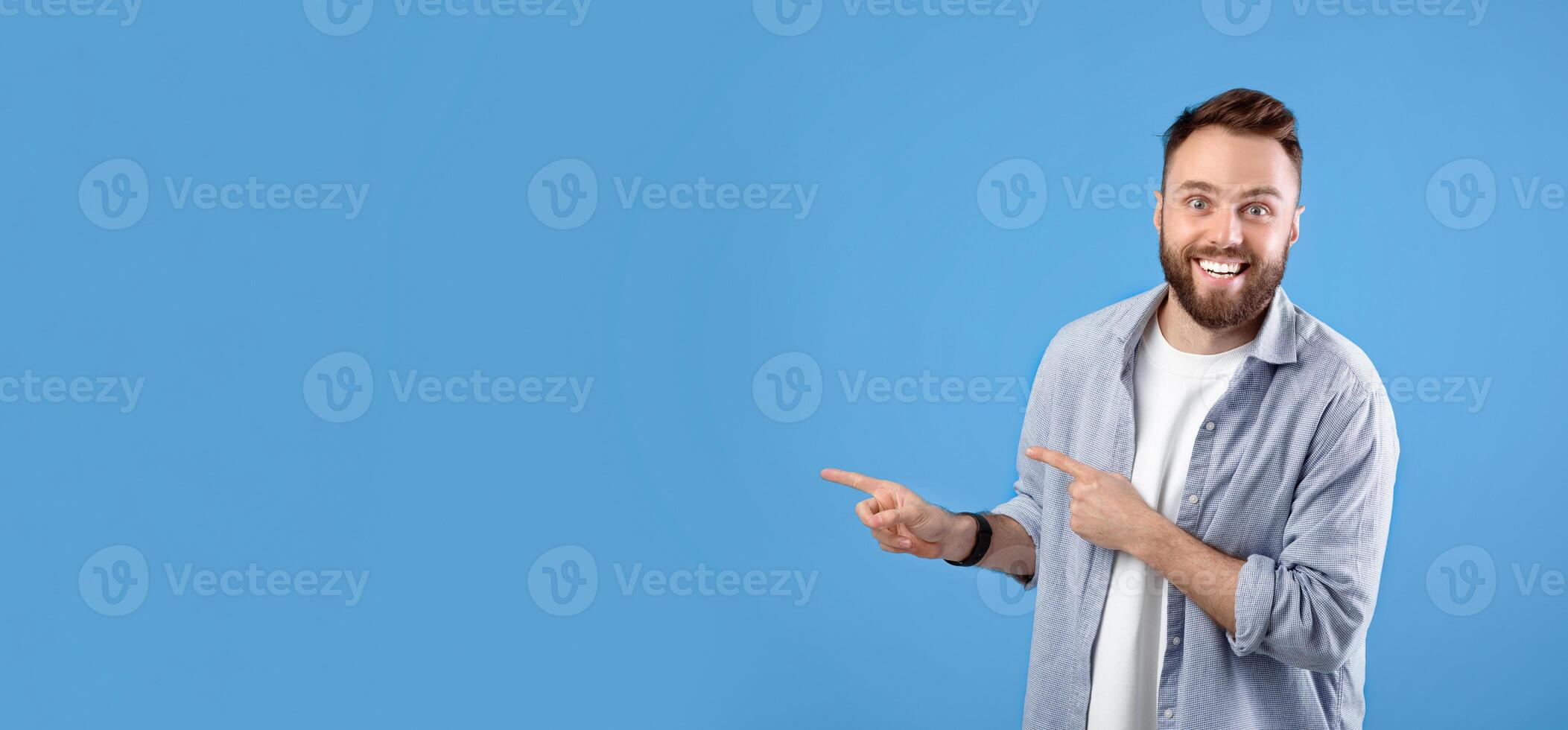 Excited young bearded guy smiling and pointing aside at empty space over blue studio background, panorama. Amazed Caucasian man having unbelievable offer, promoting product photo