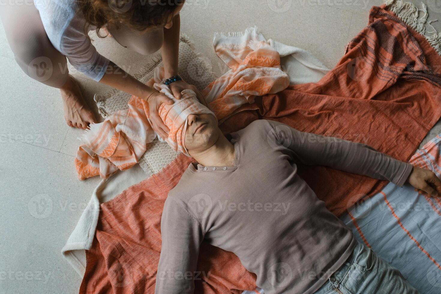 Man lying on the floor while a person ties a cloth around his eyes during a relaxation or meditation practice in a serene indoor setting. photo