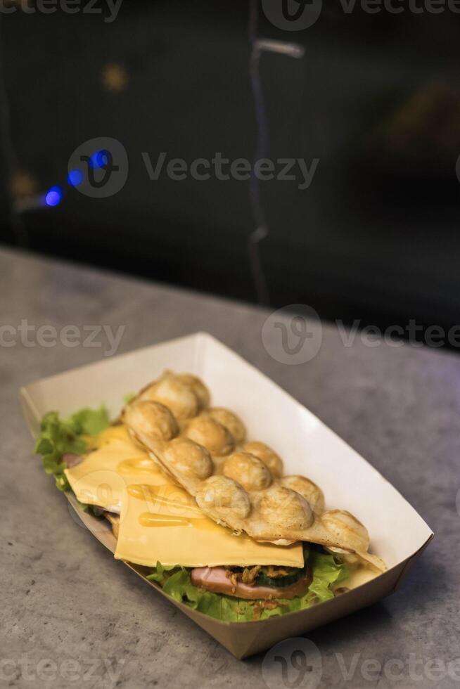 Tasty bubble waffle filled with cheese and ham served on fresh lettuce in a food market at evening hours photo