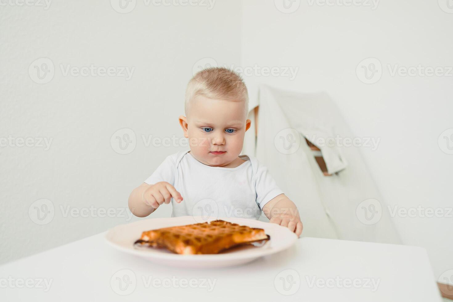 An adorable baby, full of curiosity, is examining a delicious waffle placed on a bright white table within a bright, minimalistic setting, creating a delightful scene of childhood exploration photo