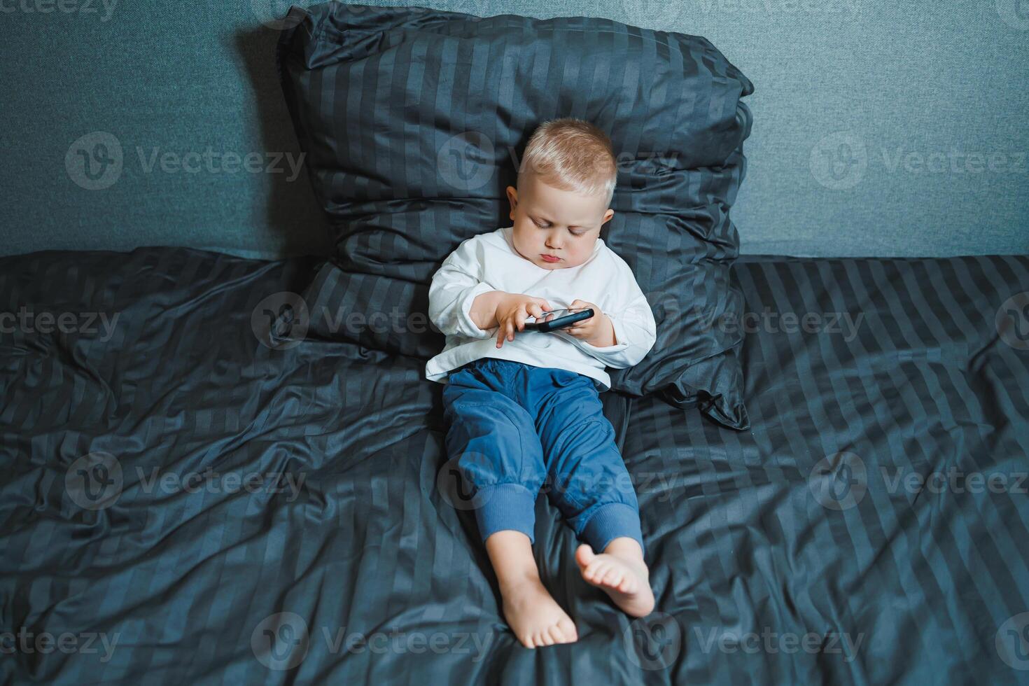 A young child can be seen fully engaged with a mobile device while comfortably relaxing on their bed, creating a delightful scene that captures the innocence and curiosity of childhood photo