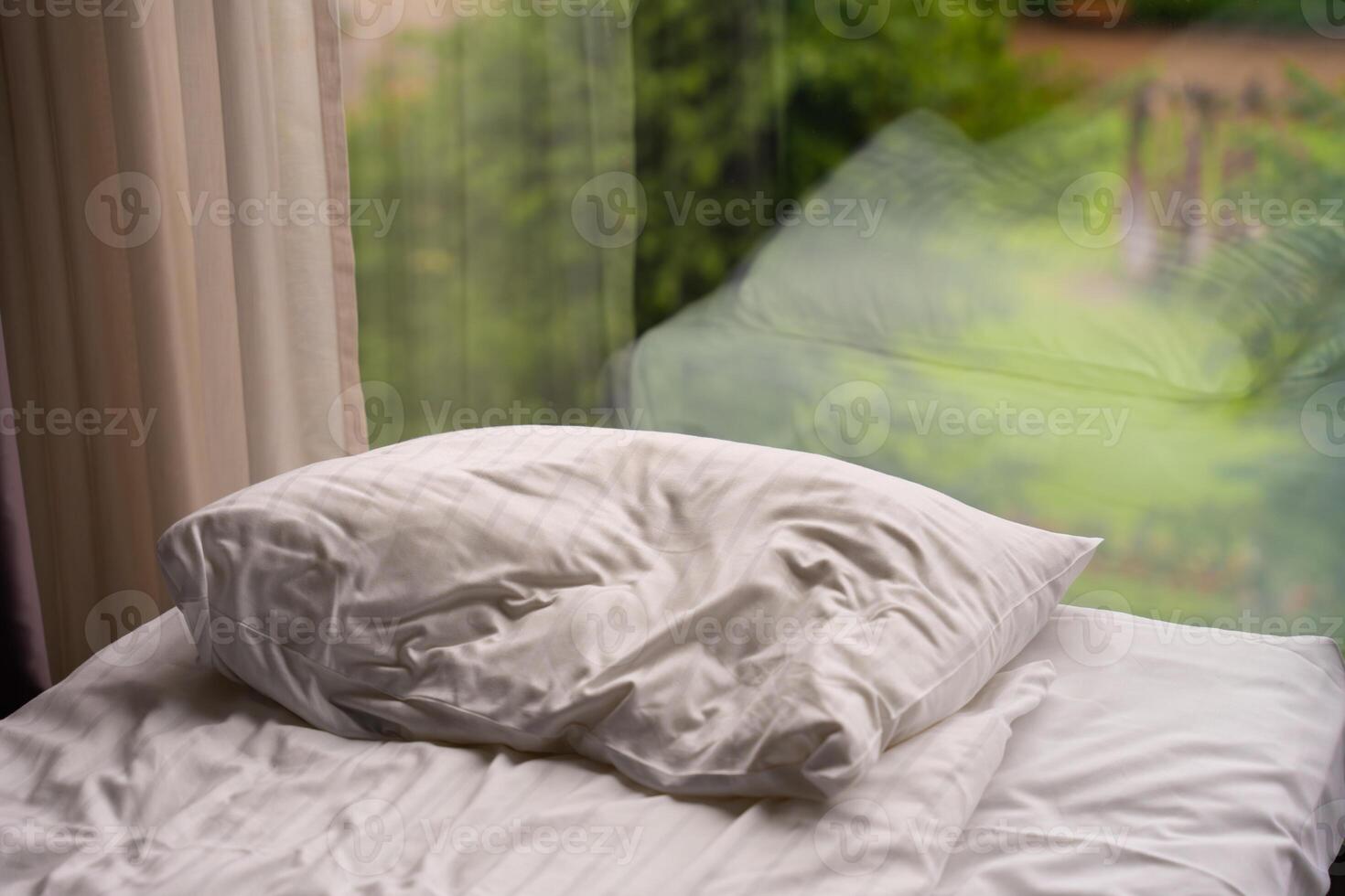White pillow on unmade bed near window with garden reflection photo