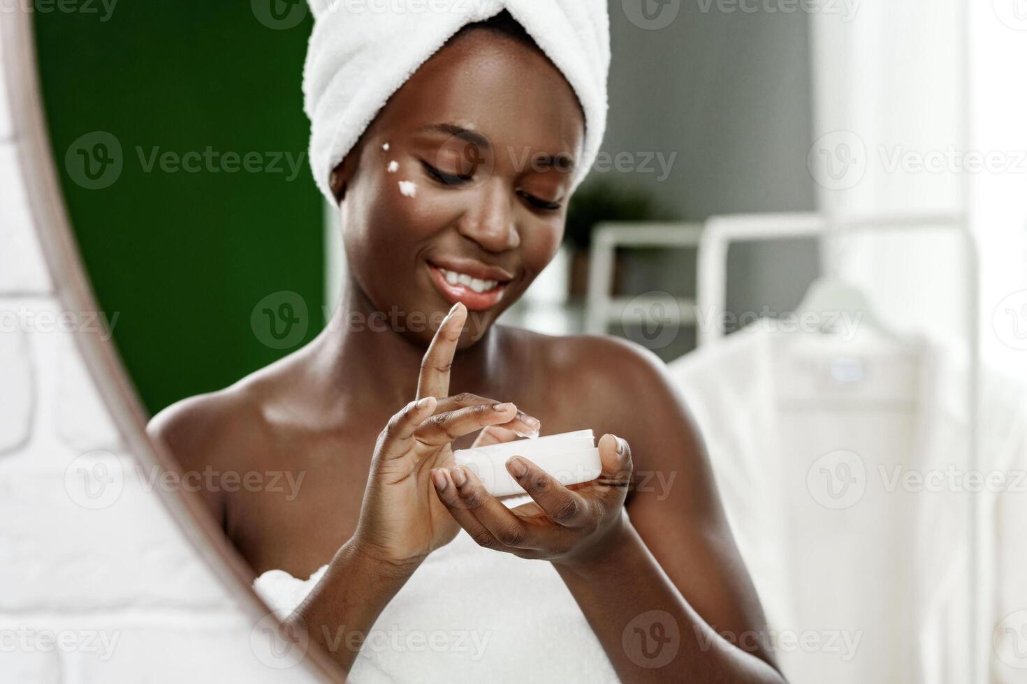 Woman applying skincare product with a towel on her head in a bright bathroom photo