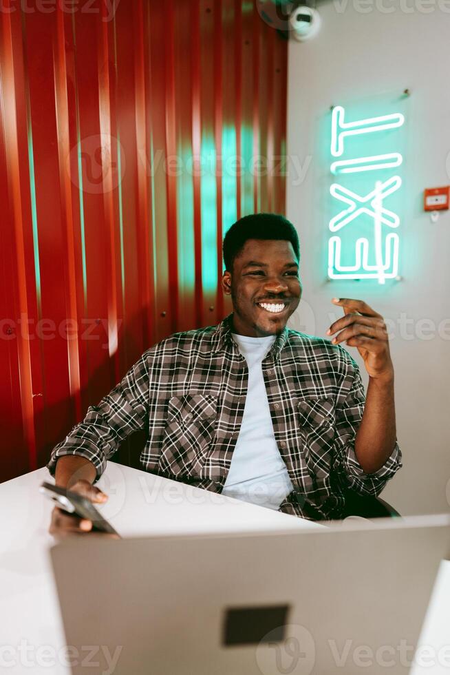 Young man engaged in work while smiling in a modern workspace with a neon exit sign in the background photo