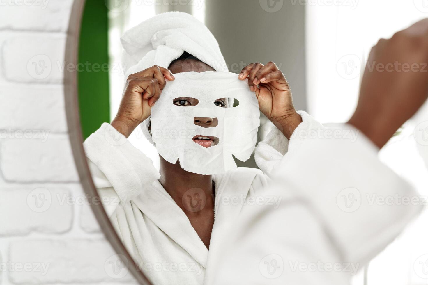 Woman applies sheet mask while looking into mirror in bright bathroom during self-care routine photo