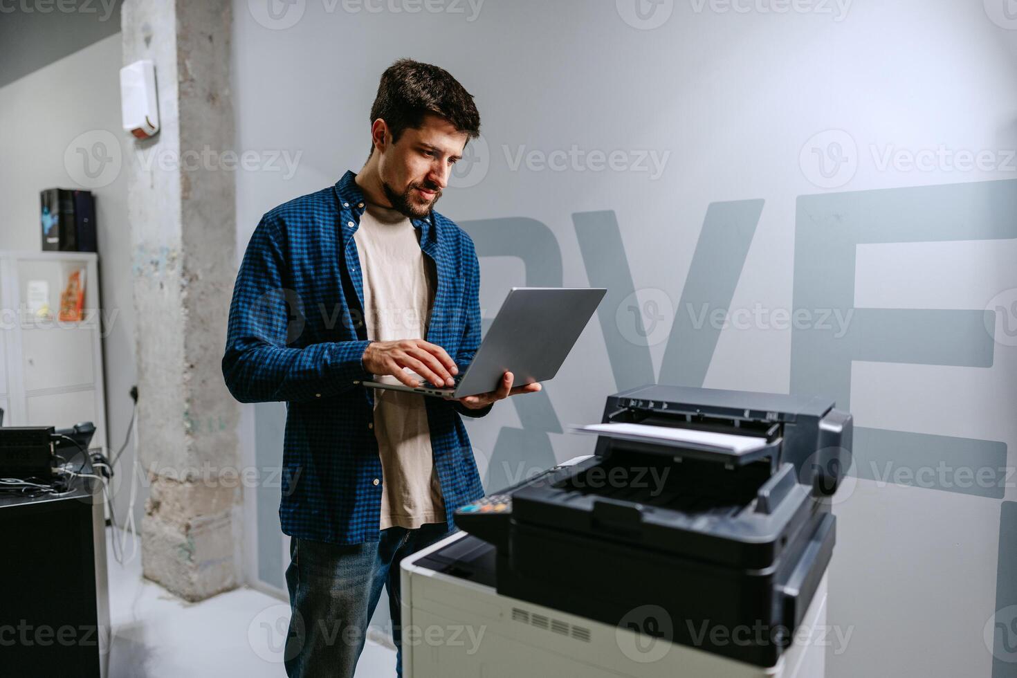 Man works on laptop next to a printer in a modern office space during the daytime photo