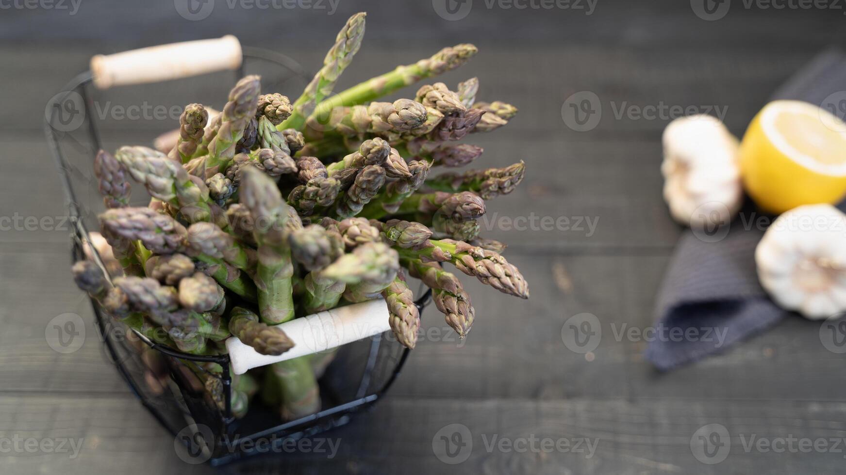 Close up of green asparagus in metal basket, garlic and lemon on a cloth, all atop a dark wooden surface photo