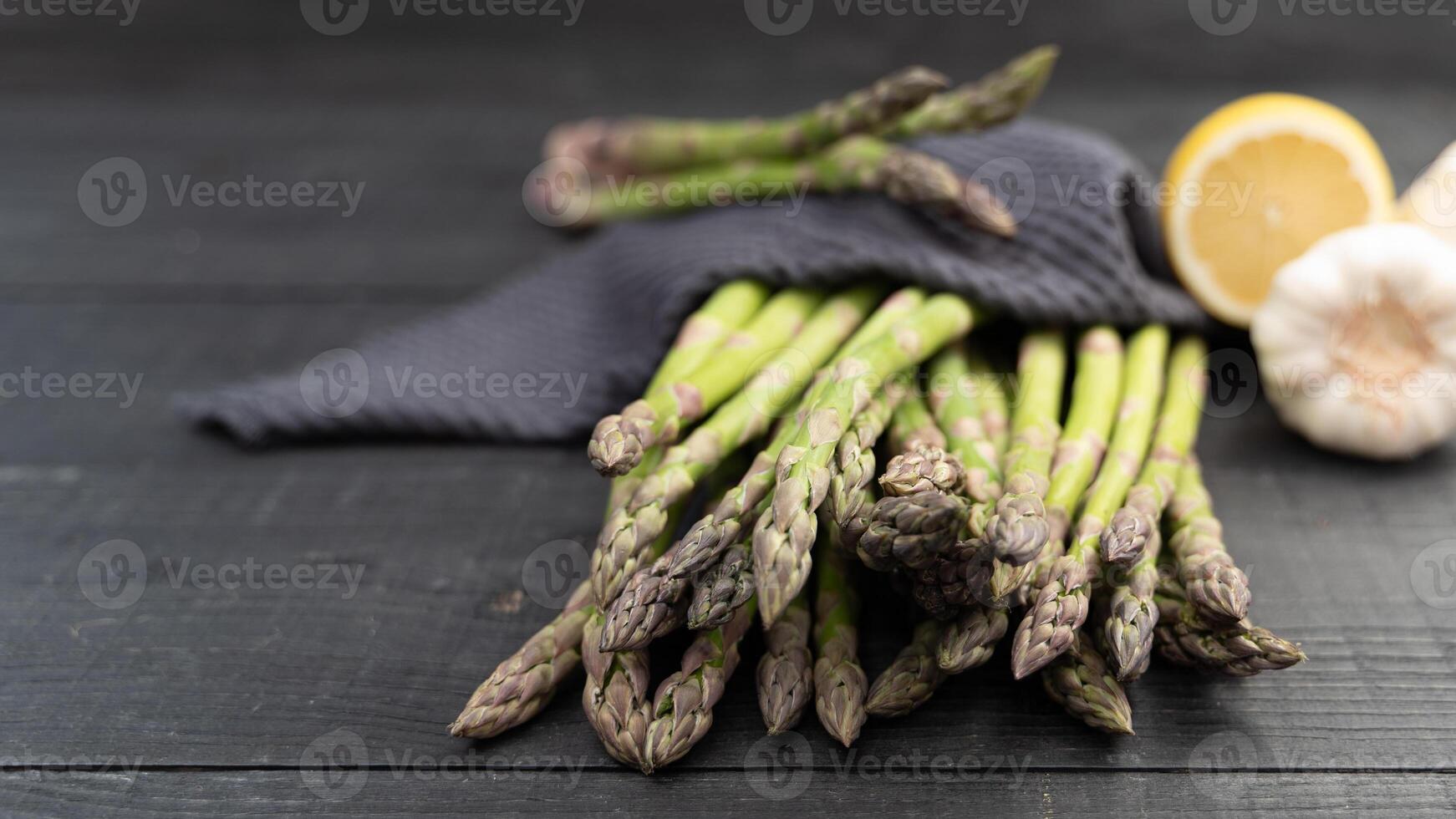 Close-up of green asparagus spears resting on dark wood with a deep blue kitchen towel, lemon and garlic in background photo