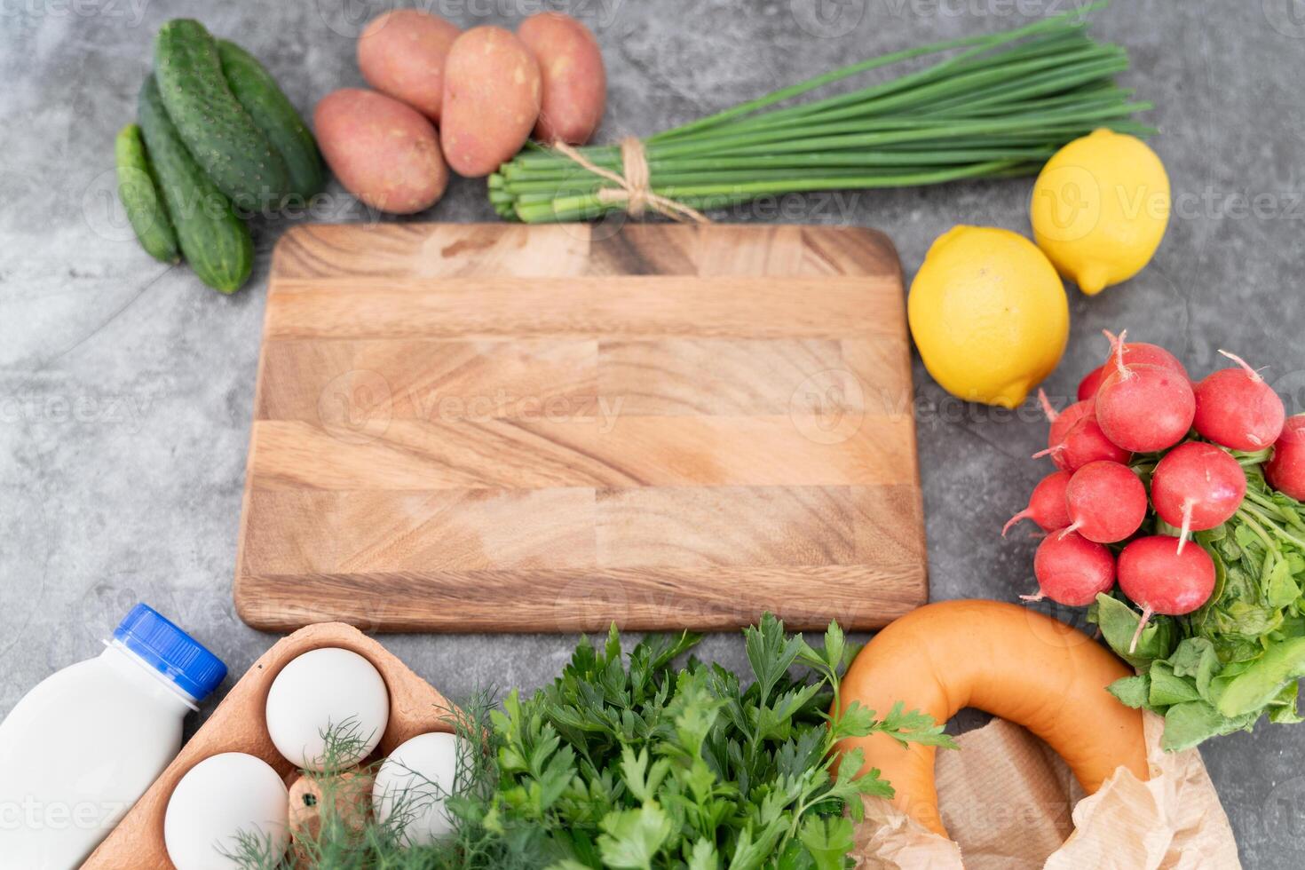 A rustic wooden board anchors the display of fresh okroshka ingredients on a cool gray background. photo