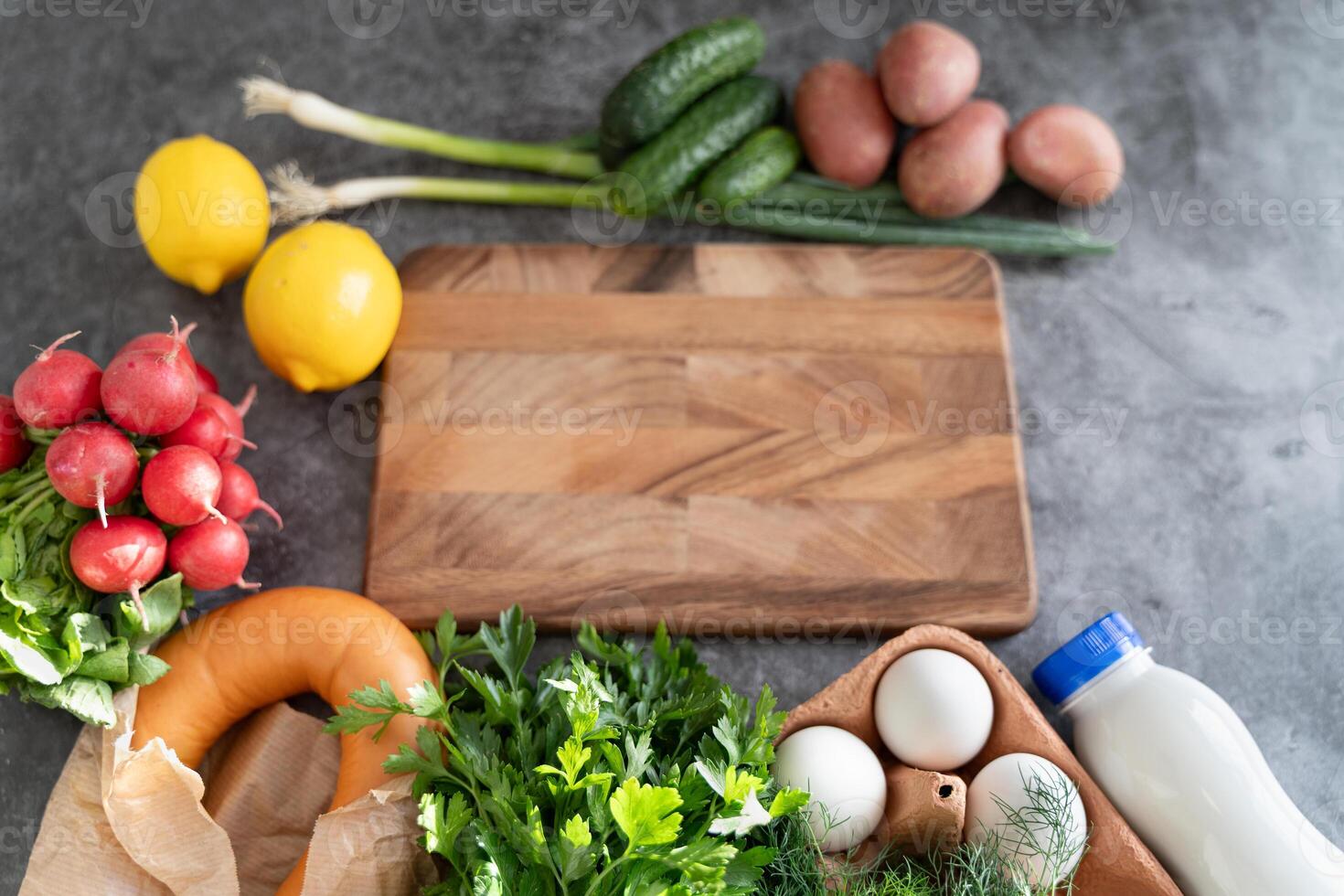 Okroshka ingredients arranged around a wooden cutting board centerpiece on a gray tabletop. photo