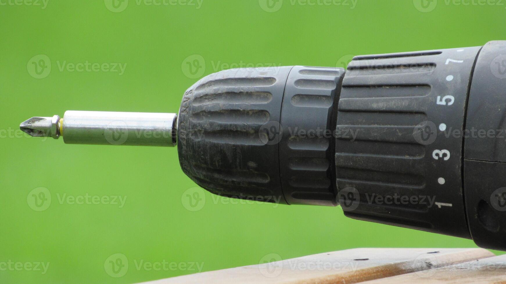 Close-up view of a black electric drill resting on a wooden surface in a green outdoor setting during daylight photo