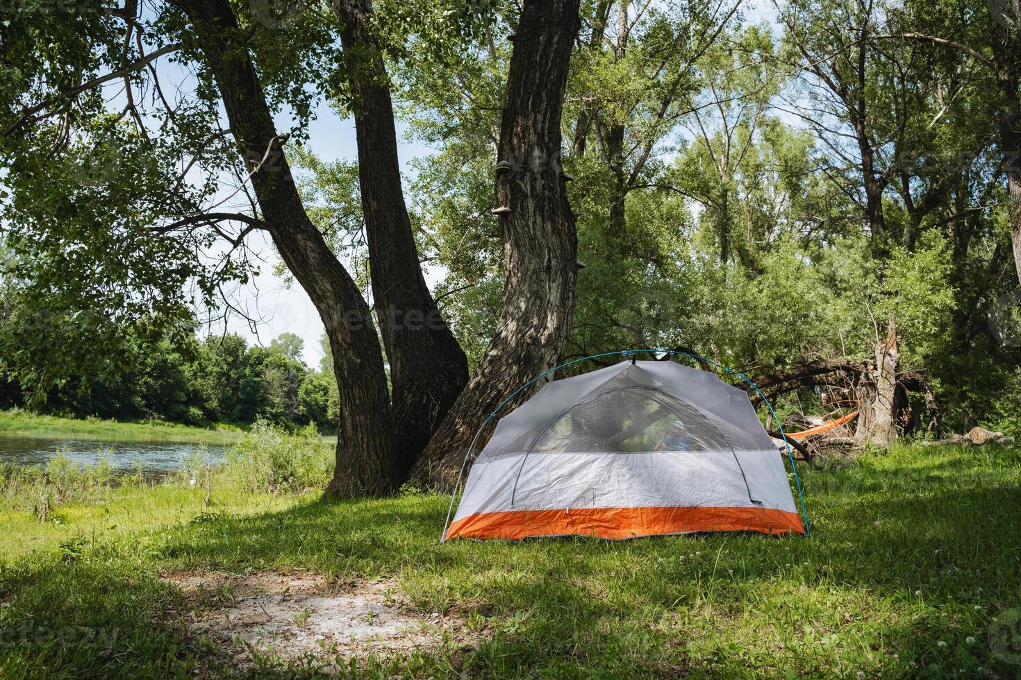 A beautiful and serene camping scene featuring a tent positioned by a tranquil riverbank under the shade of lush trees, creating the perfect spot for relaxation and leisure in natures embrace photo