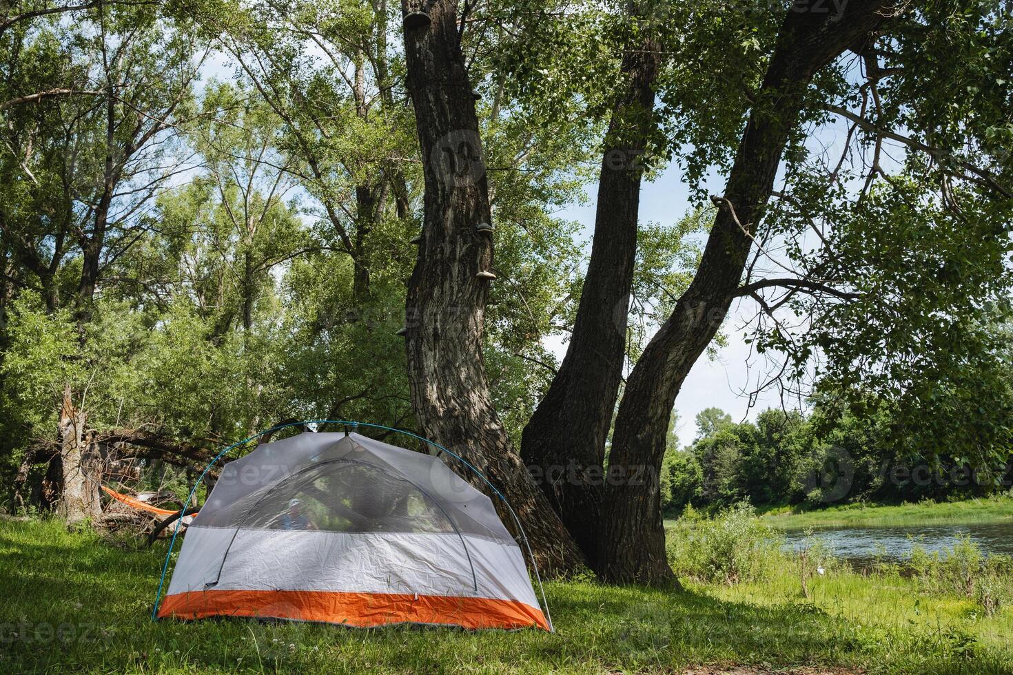 This peaceful campsite by a flowing river has a cozy tent under a lovely tree canopy, ideal for relaxation and outdoor activities in a beautiful setting with stunning scenery and lush greenery photo