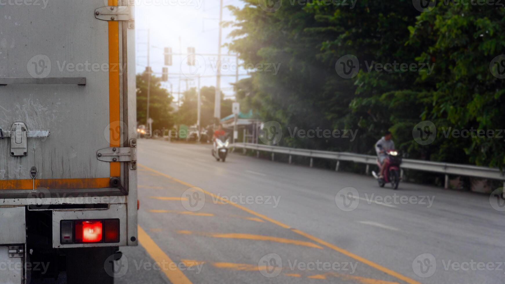 Truck car stop and turn on brake light on aspahlt road. There are yellow lines on the road. Blurred of motorcyclists on the side of the road have road barriers and trees. photo