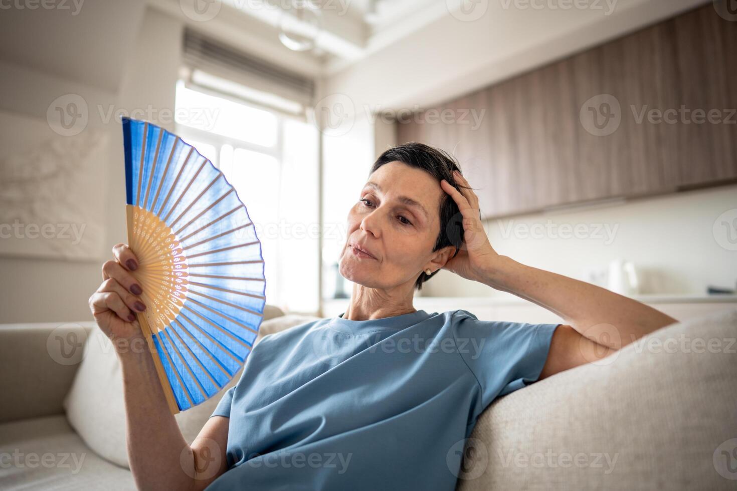 Weary older woman using fan to cool down in sweltering hot apartment. Heat exhaustion suffering photo