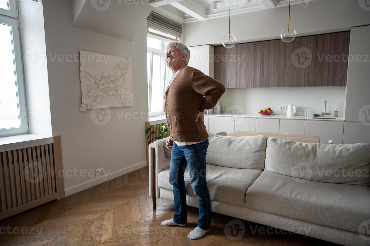 Sad elderly man painfully straightens hunched back after rising from couch with severe spinal pain photo