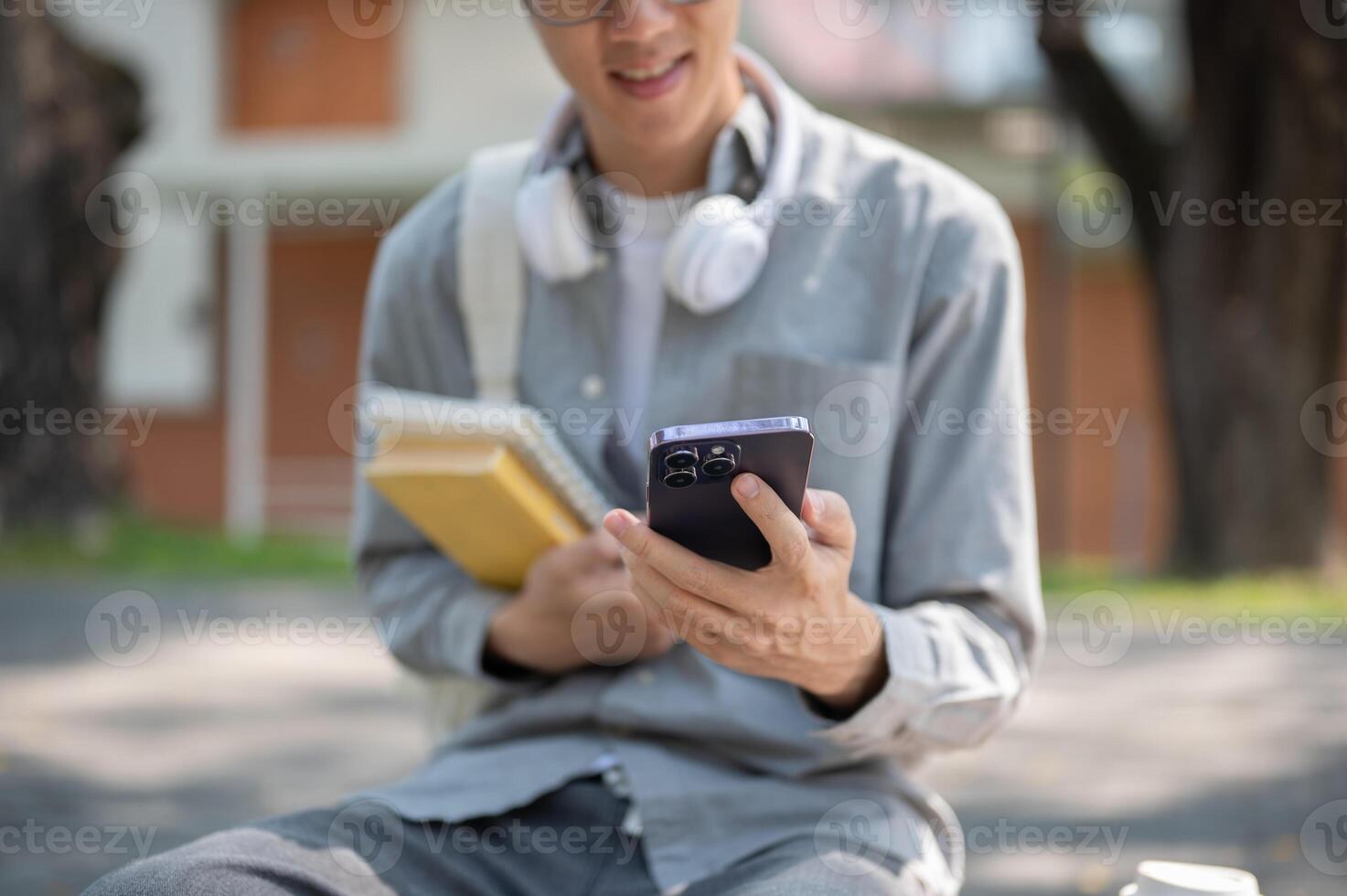 Close up of an asian man with glasses smiling while looking at his phone, sitting on sidewalk bench. photo