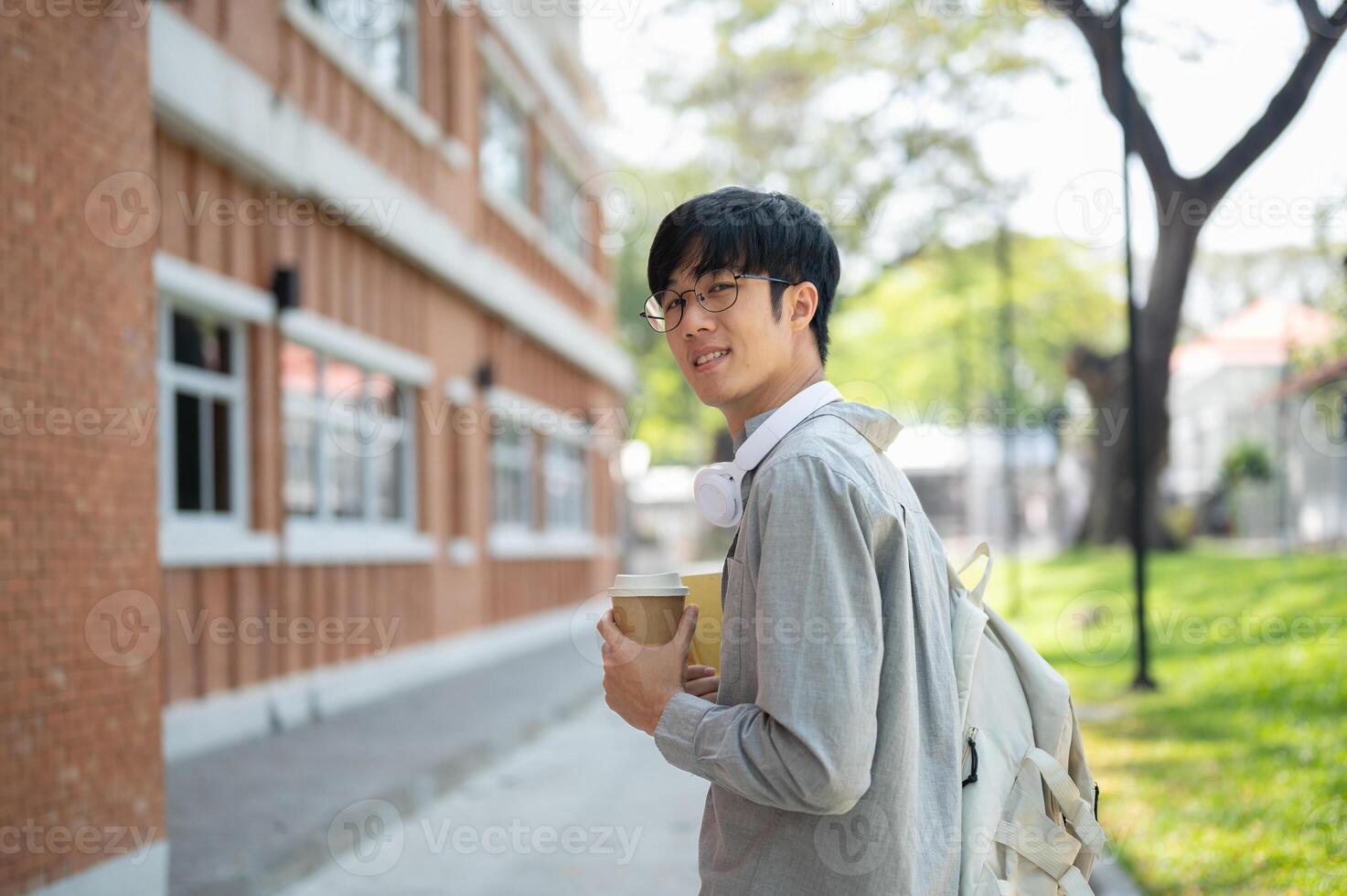 Asian man with glasses is holding a coffee cup and looking at camera while standing near brick wall photo