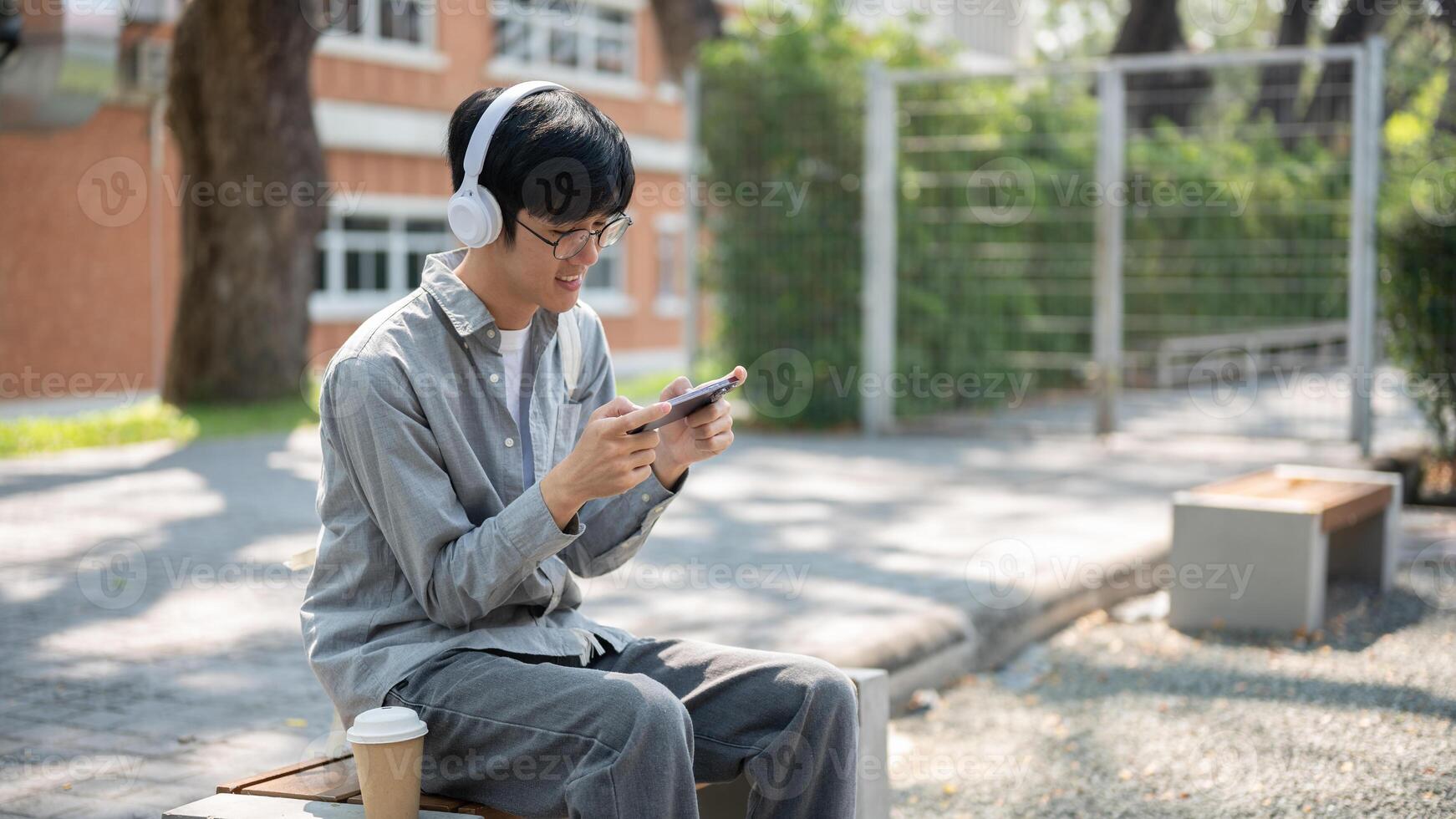 Smiling asian man with glasses is watching or playing game on his phone, sitting on the bench. photo
