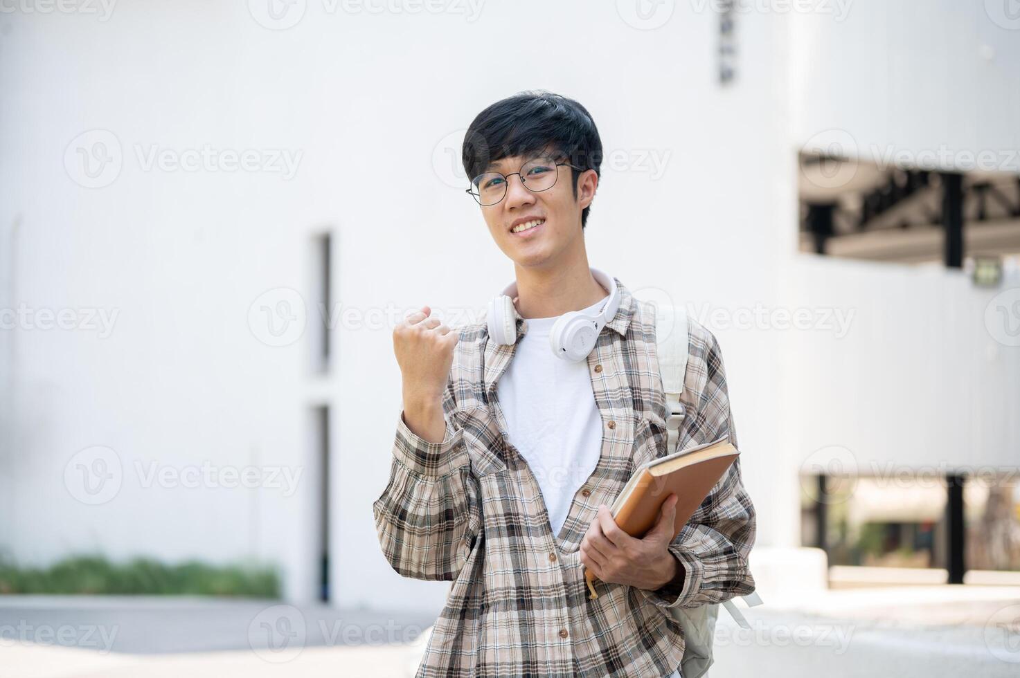 An asian man with glasses is holding books and making a fist while standing in front of a building. photo