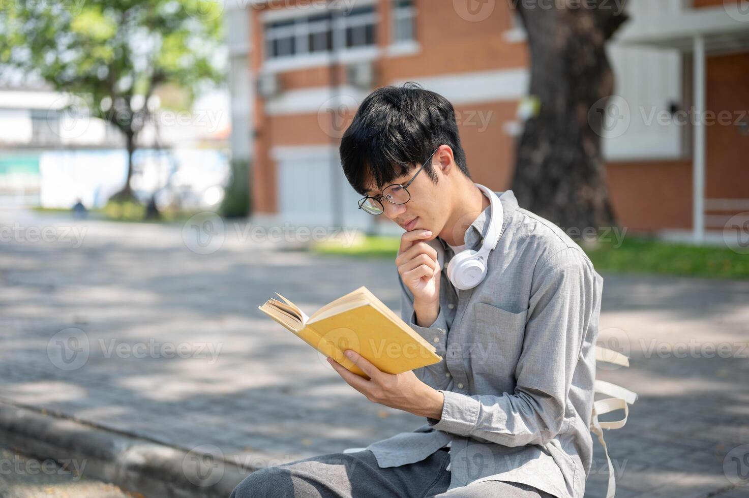 Asian man with glasses is smiling while reading a book with yellow cover, sitting on sidewalk bench. photo
