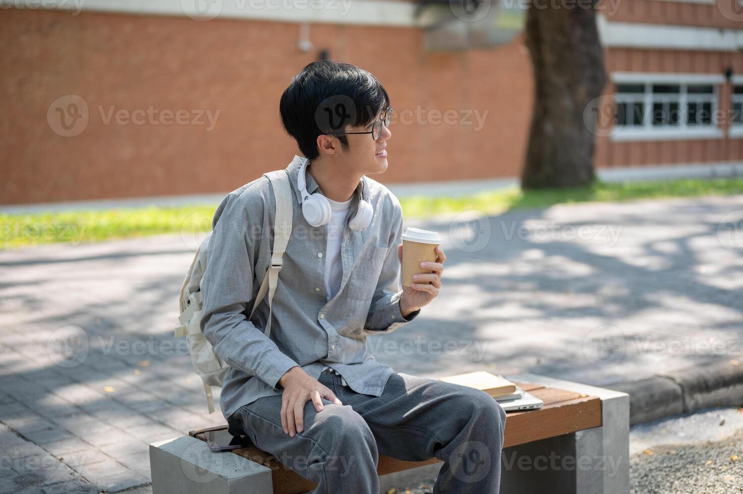 An asian man with glasses is looking away while holding a coffee cup, sitting on the sidewalk bench. photo