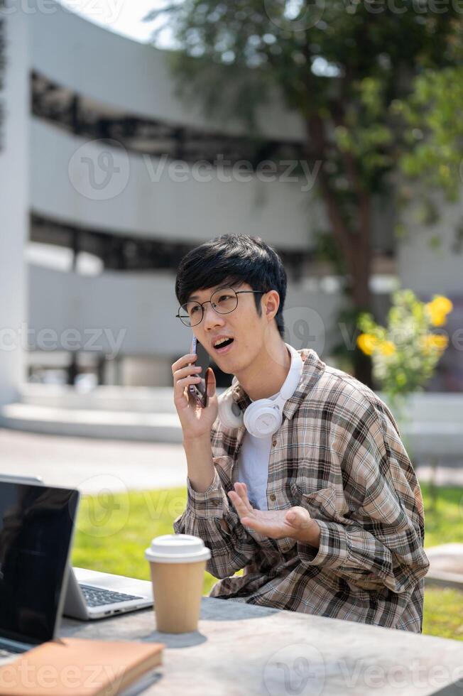 An asian man with glasses talking on phone while sitting with a laptop at a concrete table in park. photo