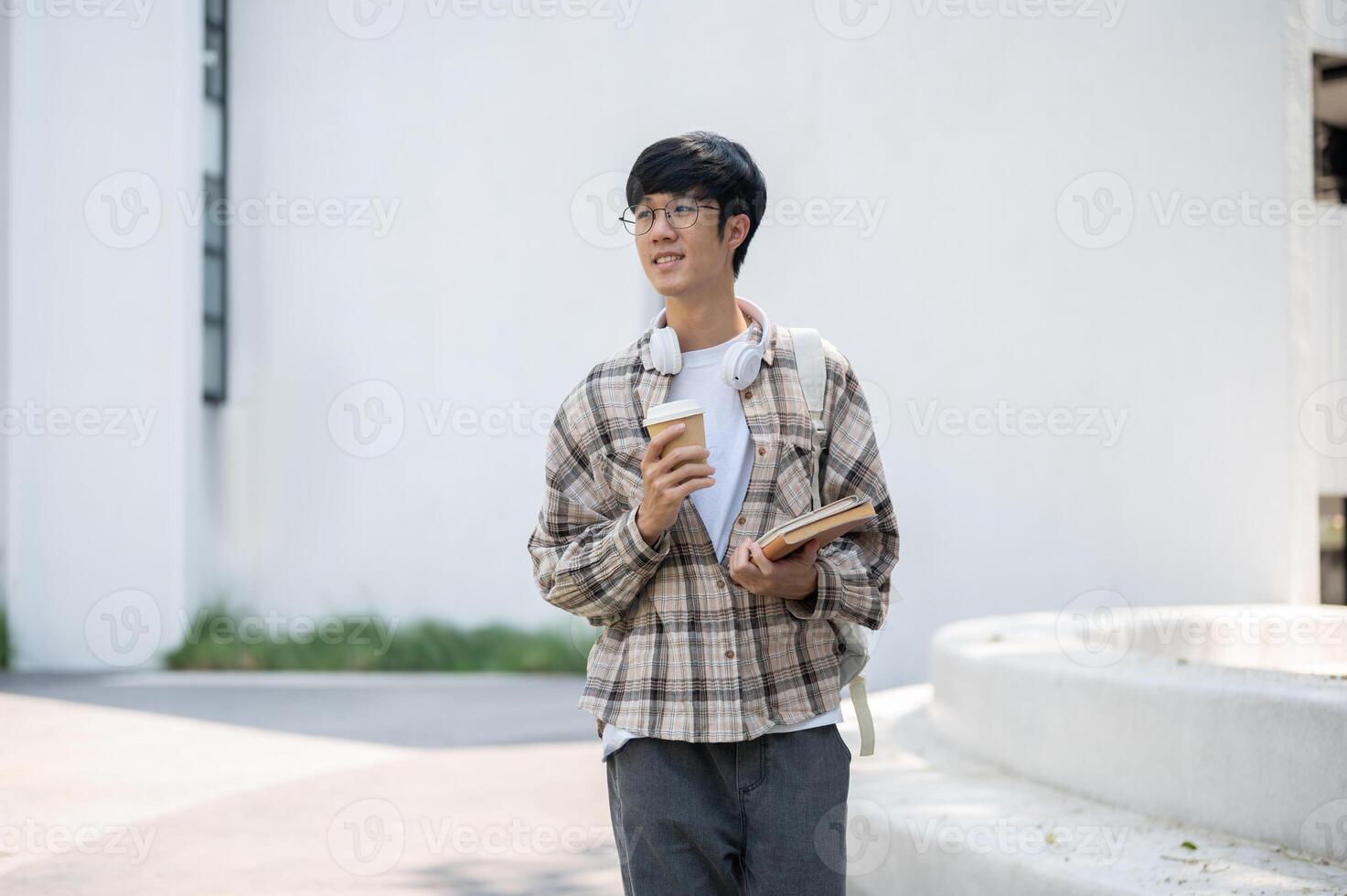 An asian man with glasses is holding coffee with books while standing or walking around the building photo