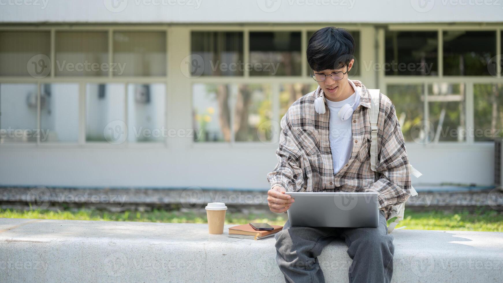 An asian man with glasses is holding laptop screen while sitting on a concrete bench in the park. photo