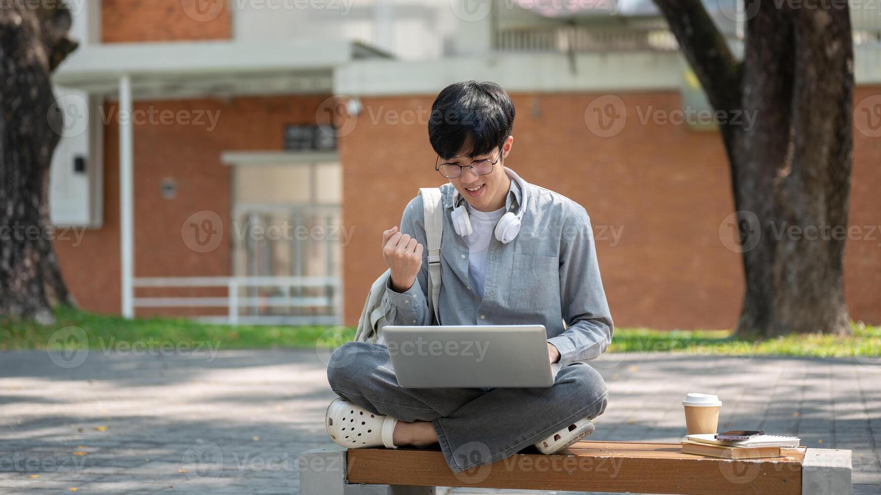 An asian man with glasses is making a fist while looking at his laptop, sitting on sidewalk bench. photo