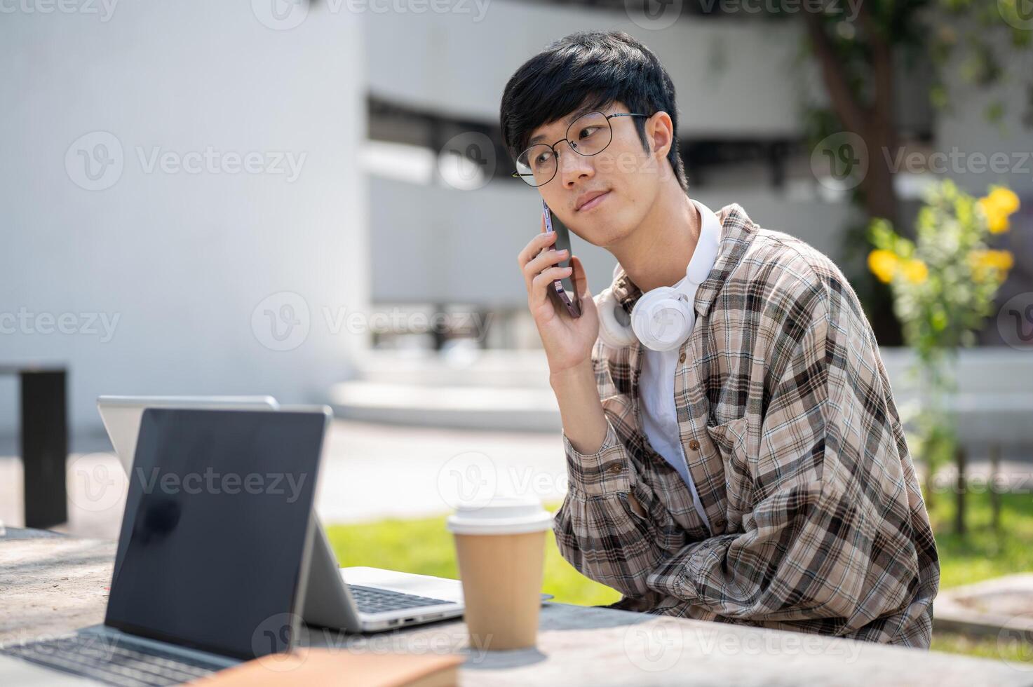An asian man with glasses is making a call while sitting with laptop at concrete table in park. photo