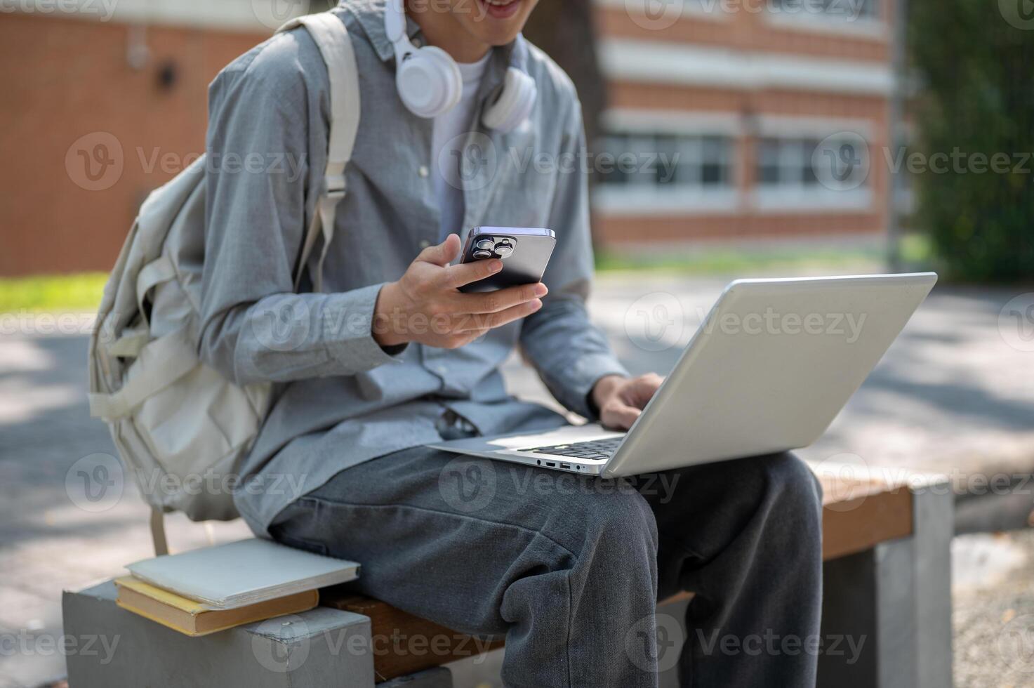 Close up of a man holding a phone while typing on laptop, sitting next with books on sidewalk bench. photo