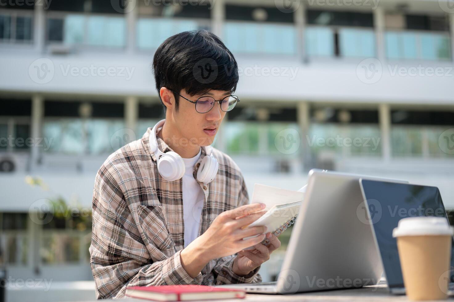 An asian man with glasses is reading a notebook while sitting with a laptop at table in park. photo