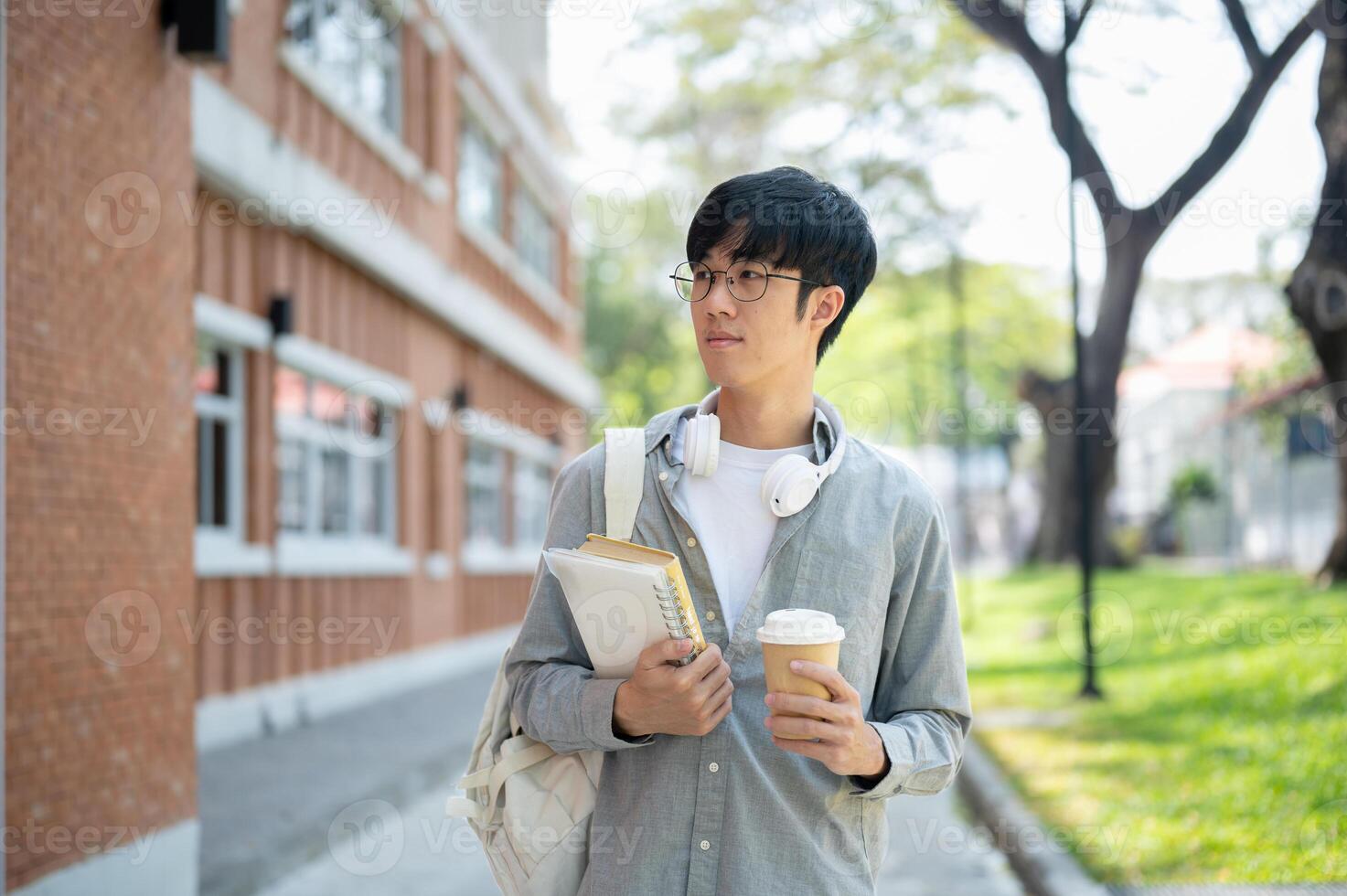 An asian man with glasses is holding books with a coffee cup while standing next to a brick wall. photo