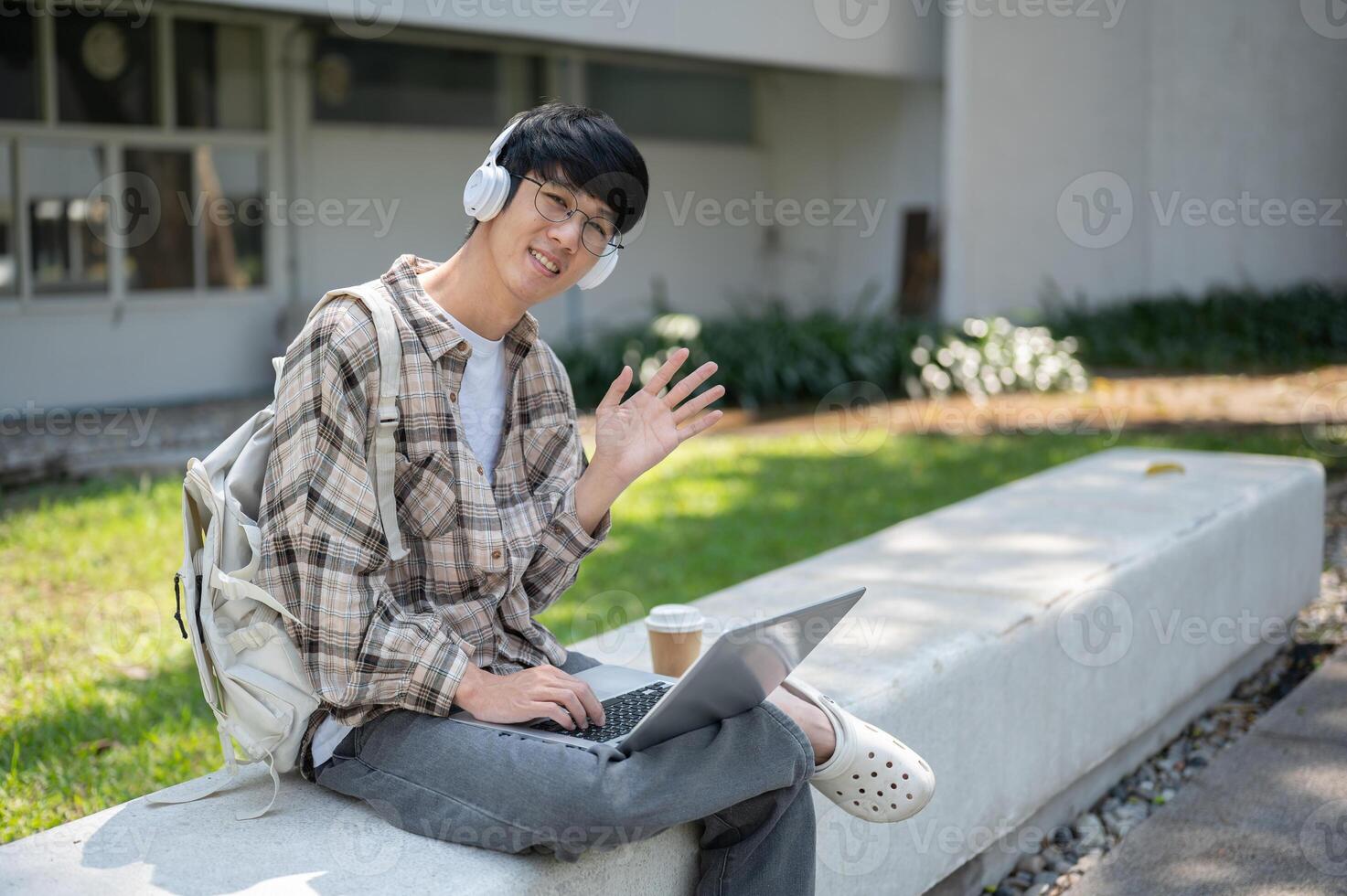 Smiling asian man with glasses and headphone waving hand while sitting with laptop on concrete bench photo