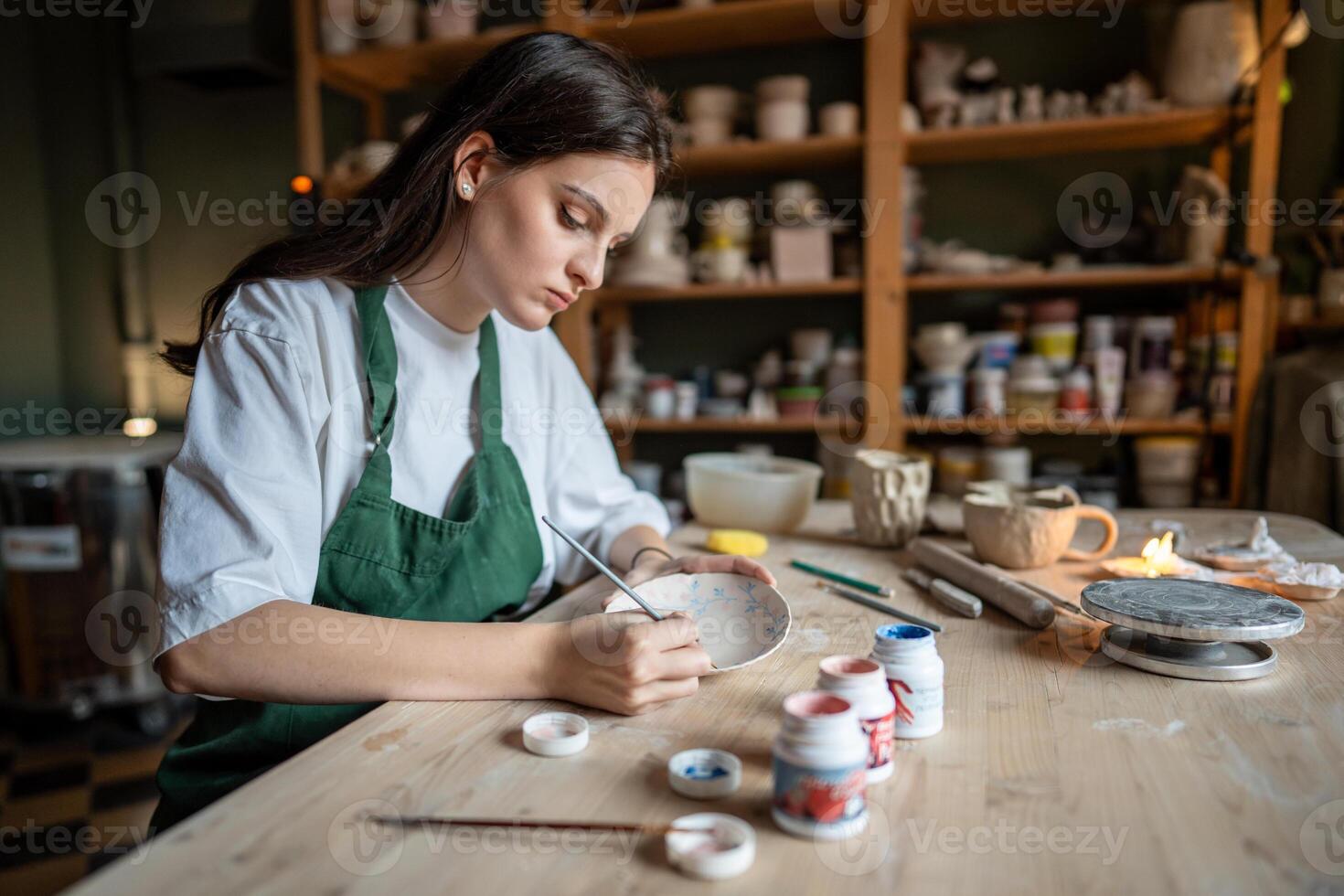 Concentrated female ceramist decorating clay tableware with paints in pottery school. Small business photo
