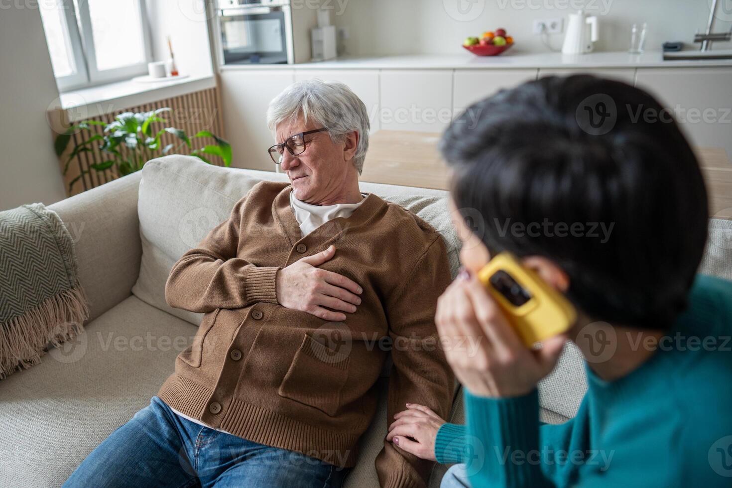 Elderly man clutching chest while sitting on chair, wife calls emergency services with phone nearby photo