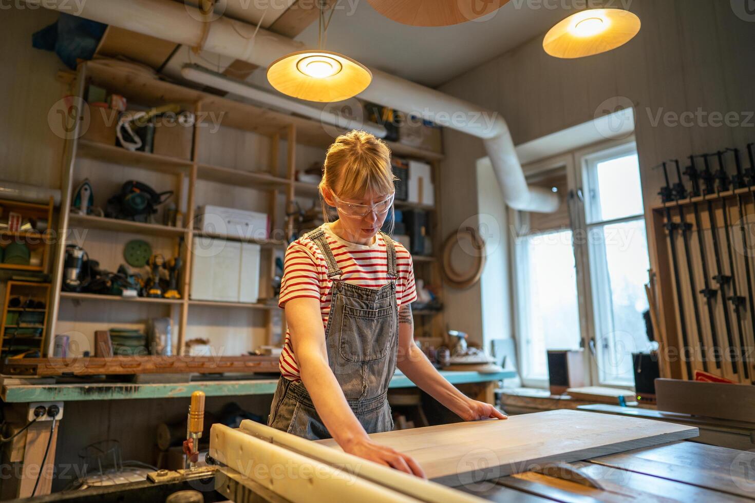 Woman carpenter in protective goggles cuts wooden piece on woodworking machine in craft workshop photo