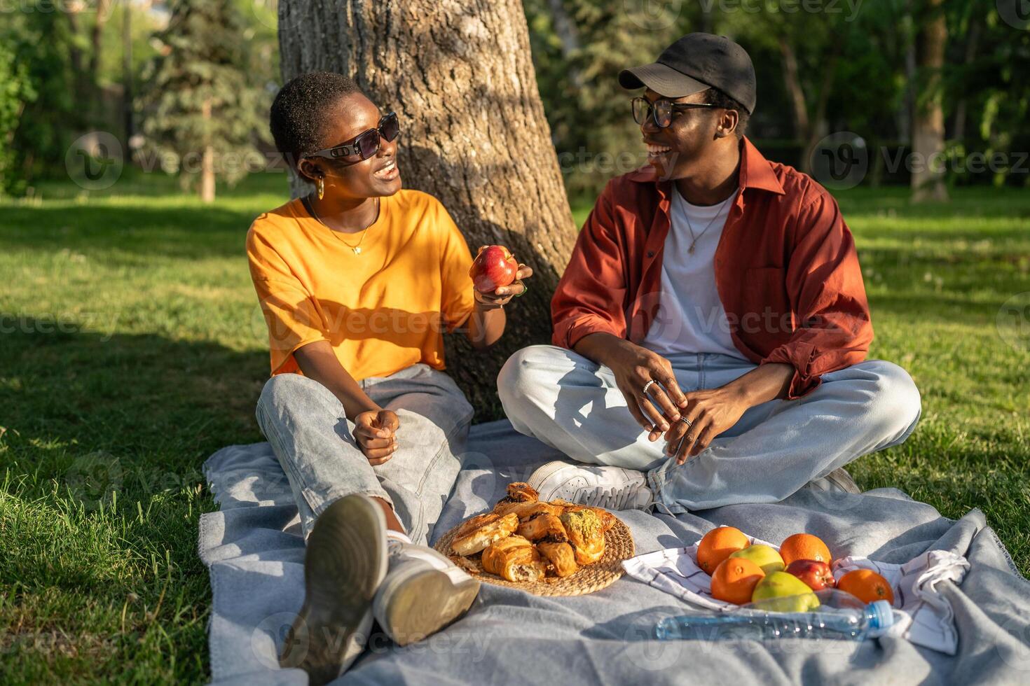 Young black couple sit on picnic blanket, eat and chat in park. Relaxed date in nature, city break photo