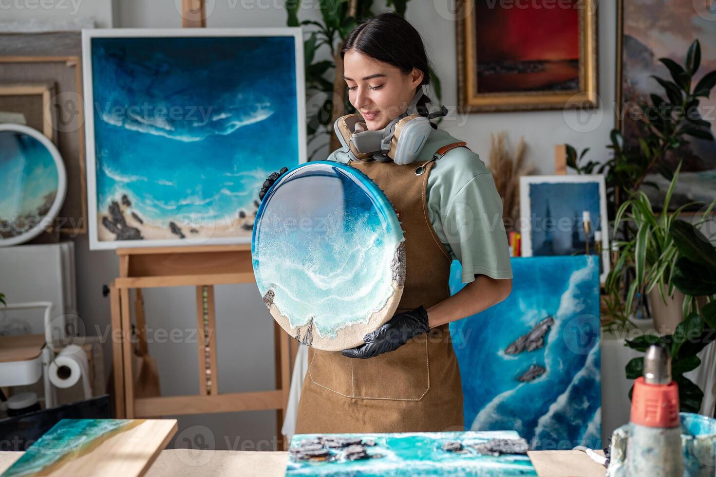Content female artist in protective gear prepares round resin artwork with ocean waves in studio photo