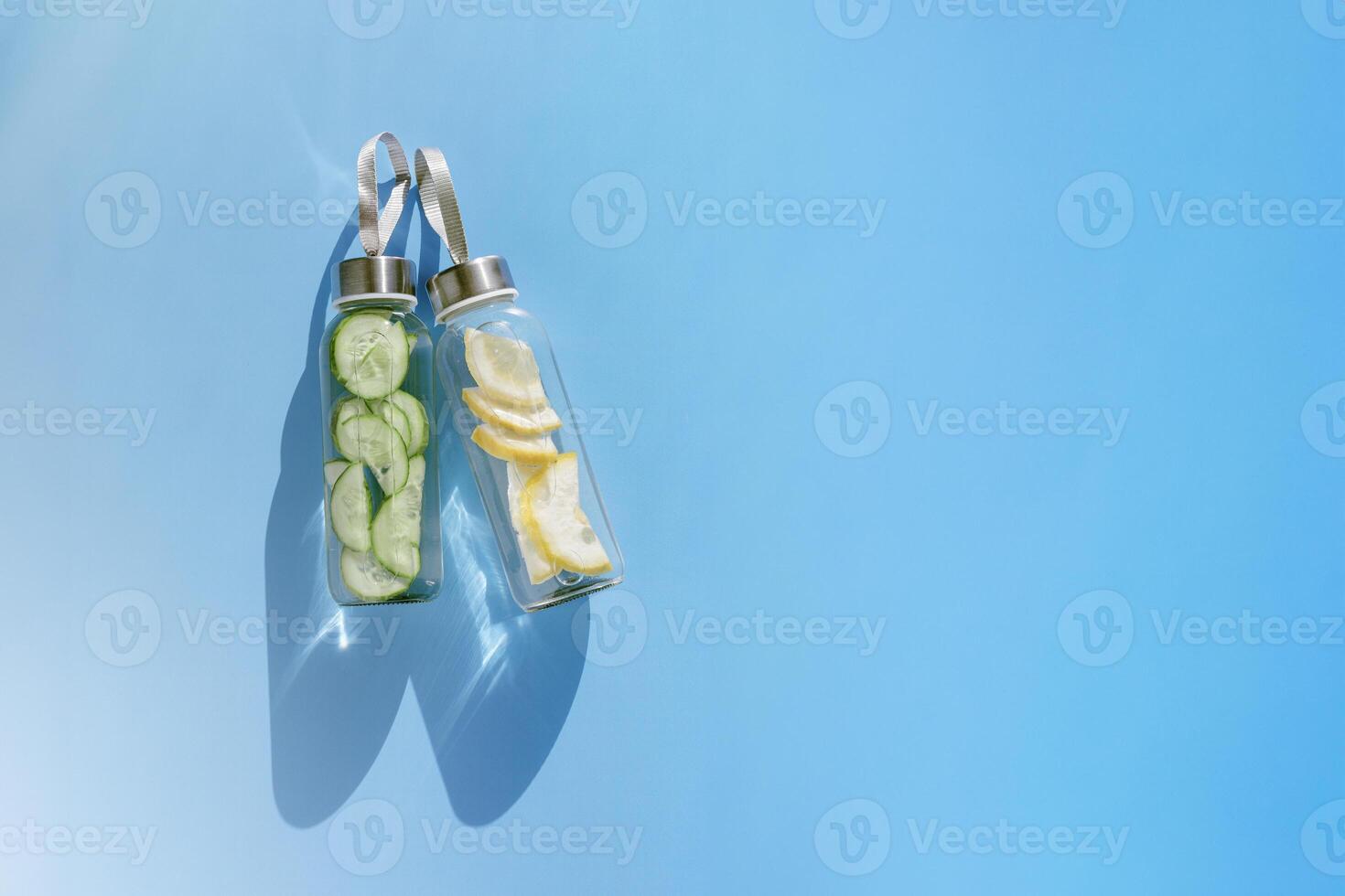 Glass bottles with lemon, cucumber water drink detox at sunlight on blue color background, copy space. Healthy infused water boost metabolism, weight loss. Aesthetic still life, refreshing drink photo
