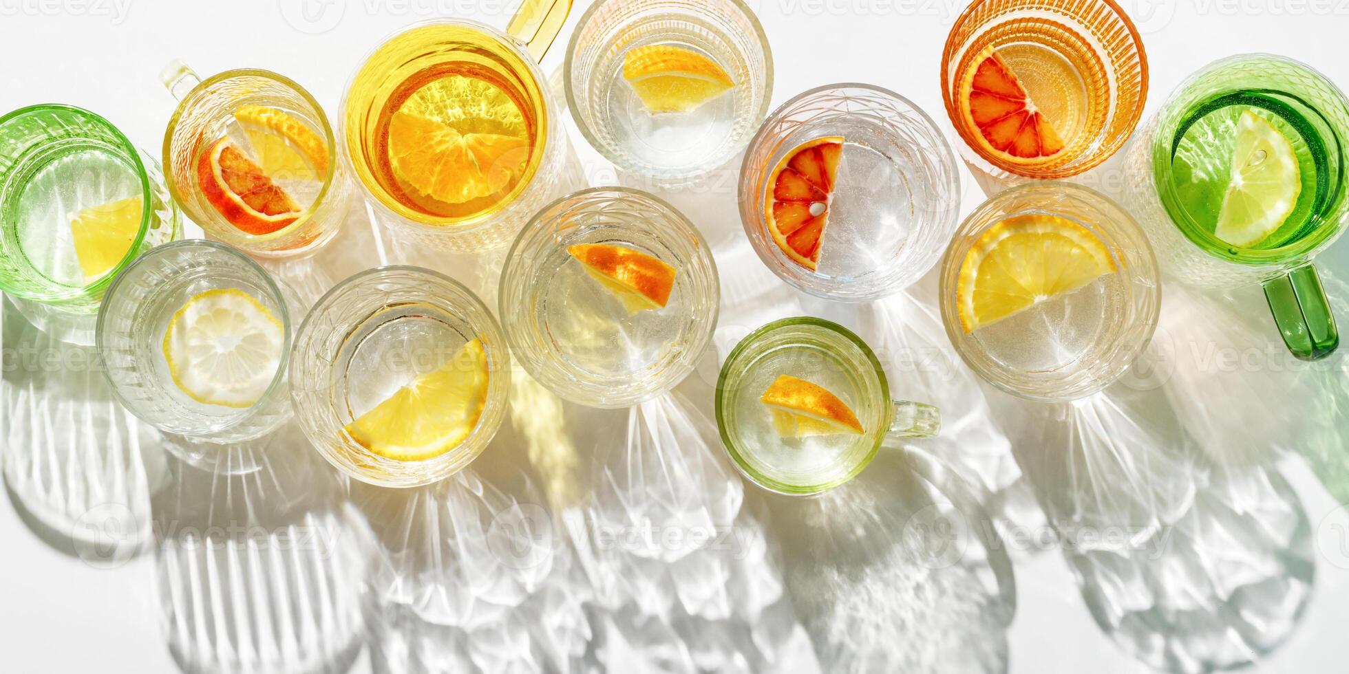 Colorful glass cups of citrus water with slices of fruit, bright glasses of detox drink with beautiful shadow at sunlight, minimal style aesthetic top view banner, creative summer drink pattern photo