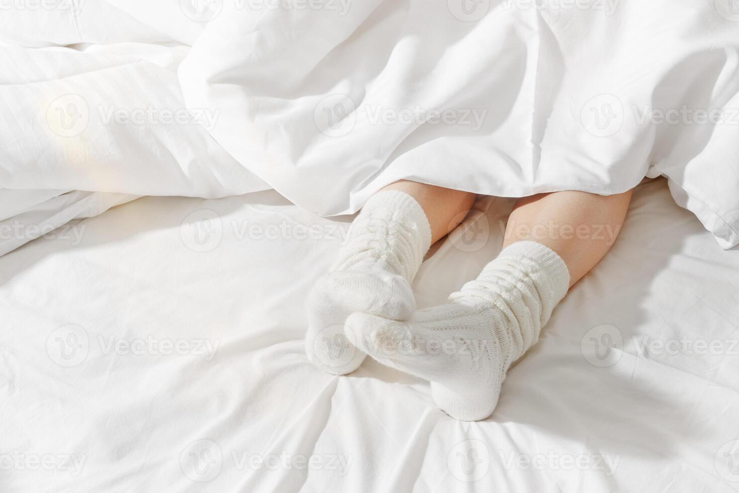 Woman wearing knitted white socks under blanket in white bed, legs close up, minimal lifestyle background with copy space, daylight. Female feet in woolen and cozy socks. Home relaxing concept. photo